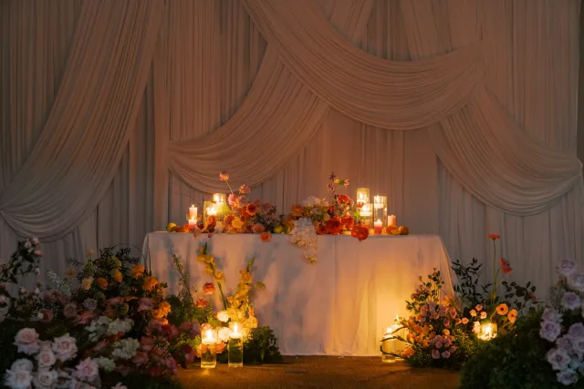 Table d’honneur de mariage fleurie devant un drapé, éclairée par des bougies
